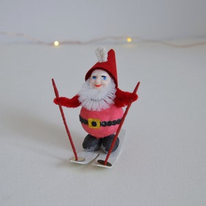 May include: A red and white Christmas gnome wearing a red hat with a white pom-pom, holding red ski poles and standing on silver skis.