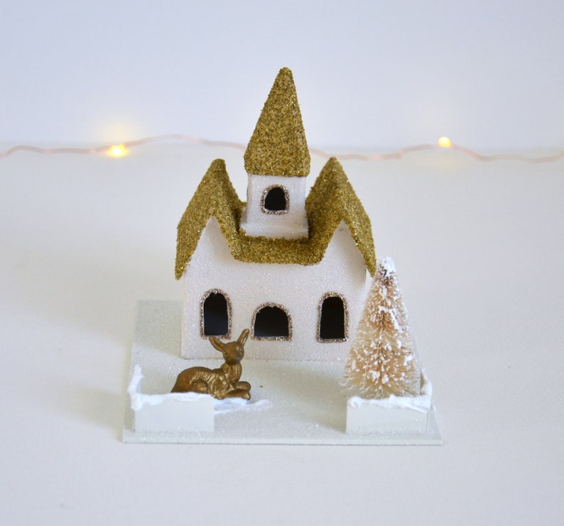Puede incluir: Una iglesia de pueblo de Navidad de papel mach&eacute; blanca y dorada con un techo de purpurina dorada, una peque&ntilde;a figura de ciervo dorada y un &aacute;rbol de Navidad blanco flocado.