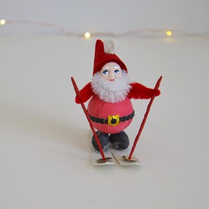 Puede incluir: Una figura de Papá Noel roja y blanca en esquís. La figura tiene una barba blanca y un gorro rojo con un pompón blanco. Los esquís son blancos con puntas rojas.