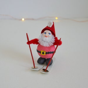 May include: A red and white Christmas ornament depicting a Santa Claus figure on skis. The figure has a white beard and a red hat with a white pom-pom. The skis are white and the ski poles are red.