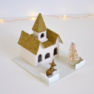 Puede incluir: Una escena de pueblo navide&ntilde;o de papel mach&eacute; con una iglesia, un &aacute;rbol peque&ntilde;o y una figura de reno dorada. La iglesia tiene un techo de purpurina dorada y paredes blancas con ventanas. El &aacute;rbol es un peque&ntilde;o &aacute;rbol de cepillo de botella blanco. El reno est&aacute; de pie sobre una base blanca.