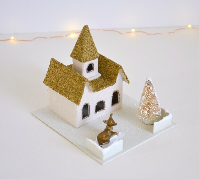 Puede incluir: Una escena de pueblo navide&ntilde;o de papel mach&eacute; con una iglesia, un &aacute;rbol peque&ntilde;o y una figura de reno dorada. La iglesia tiene un techo de purpurina dorada y paredes blancas con ventanas. El &aacute;rbol es un peque&ntilde;o &aacute;rbol de cepillo de botella blanco. El reno est&aacute; de pie sobre una base blanca.