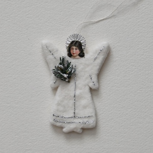 Può includere: Ornamento natalizio a forma di angelo in feltro bianco, con volto dipinto, alone argentato e dettagli glitterati. L'angelo tiene un piccolo rametto e ha un filo per appenderlo. Una decorazione festiva.