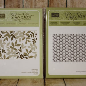 May include: Two Big Shot die-cut embossing folders. One features a leaf design in shades of brown and tan. The other has a geometric honeycomb pattern in shades of gray. Both are in green packaging.