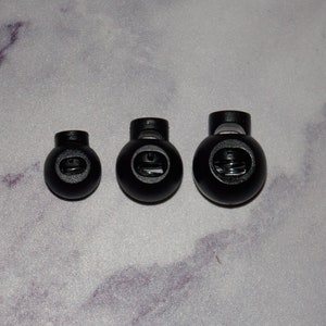 May include: Three black plastic toggle stoppers of different sizes. The stoppers have a spring inside and are used for securing cords or straps.