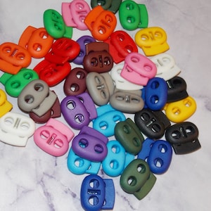May include: Assortment of colorful plastic toggle clasps, also known as cord locks, in various colors including red, blue, green, yellow, purple, brown, white, black, and gray.