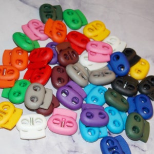 May include: A collection of colorful plastic toggle clasps, also known as cord locks, in various colors including red, green, blue, yellow, orange, purple, brown, and gray. These clasps are used for adjusting the length of cords, straps, and shoelaces.
