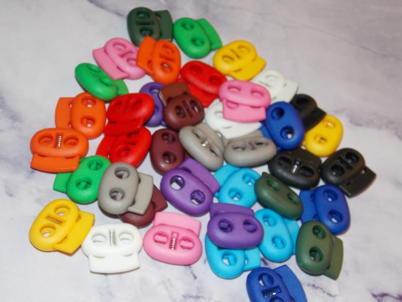 May include: A collection of colorful plastic toggle clasps, also known as cord locks, in various colors including red, green, blue, yellow, orange, purple, brown, and gray. These clasps are used for adjusting the length of cords, straps, and shoelaces.