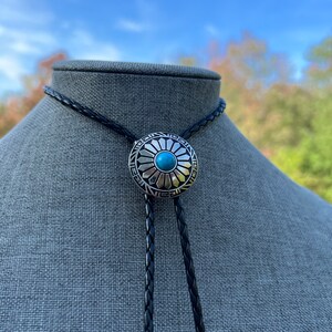 Pode incluir: Um cord&atilde;o de bolo de couro preto com um deslizador de bolo de prata e turquesa com um design circular com uma pedra turquesa no centro.