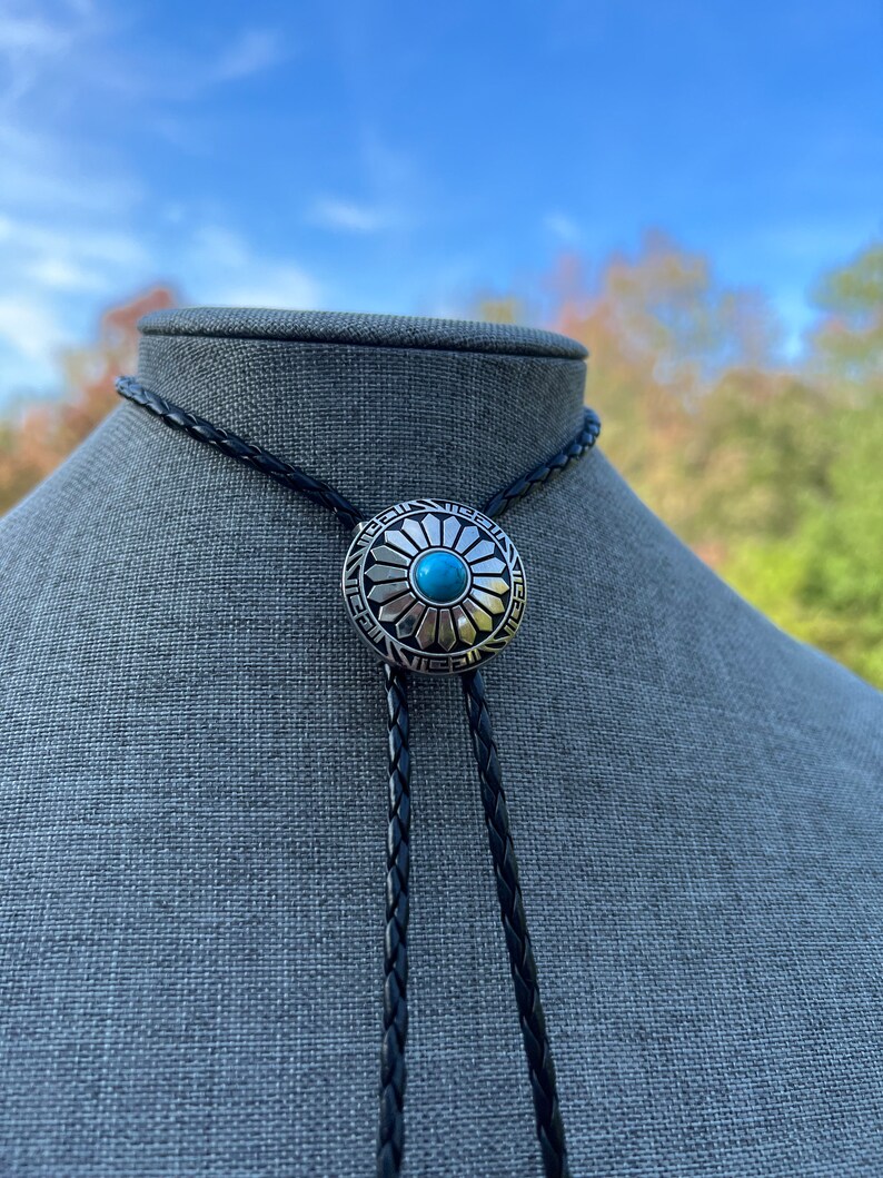 Pode incluir: Um cord&atilde;o de bolo de couro preto com um deslizador de bolo de prata e turquesa com um design circular com uma pedra turquesa no centro.