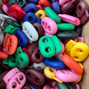 May include: A large assortment of colorful plastic toggle clasps. The clasps are in various colors, including red, blue, green, yellow, brown, and pink. They are all oval-shaped with two holes in the center.