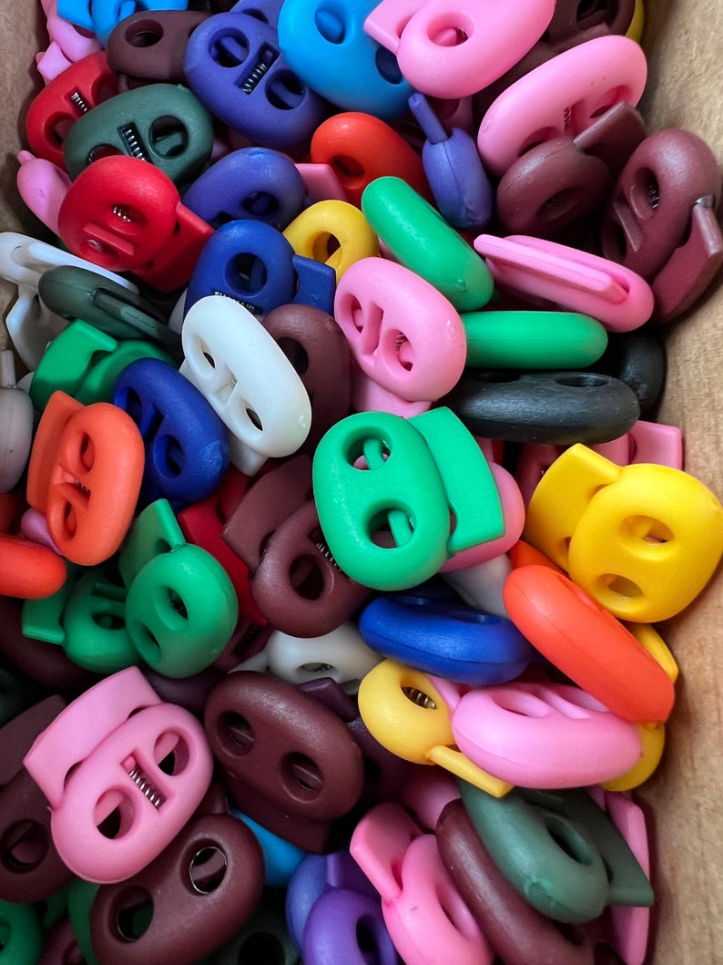 May include: A large assortment of colorful plastic toggle clasps. The clasps are in various colors, including red, blue, green, yellow, brown, and pink. They are all oval-shaped with two holes in the center.