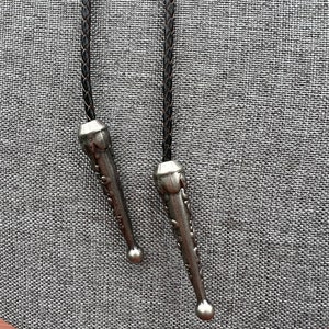 Op de afbeelding: Twee zilveren bolo tie-uiteinden met ingewikkelde ontwerpen, bevestigd aan bruine leren koorden.