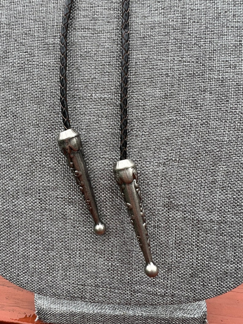 Op de afbeelding: Twee zilveren bolo tie-uiteinden met ingewikkelde ontwerpen, bevestigd aan bruine leren koorden.