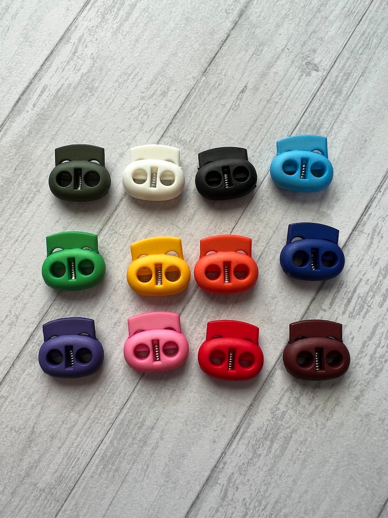 May include: Twelve plastic toggle cord locks in various colors, including green, white, black, blue, orange, red, pink, purple, brown, and yellow. The cord locks have two holes and a spring mechanism.