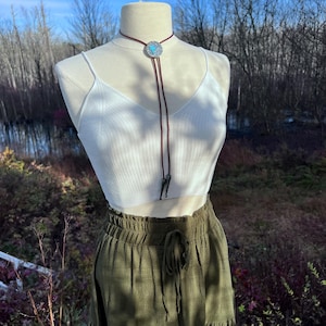 May include: A white crop top with a brown bolo tie featuring a silver and turquoise pendant. The model is wearing a green skirt with a drawstring waist.