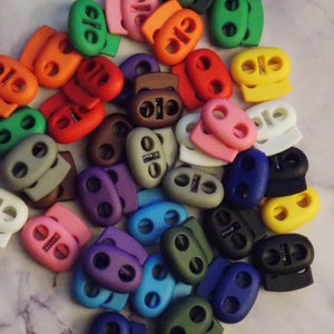 May include: A variety of colorful plastic toggle clasps, also known as cord locks, in various colors including red, blue, green, yellow, purple, gray, brown, and white.