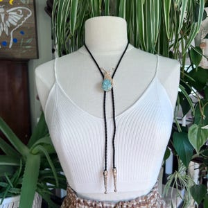 May include: A bolo tie with a black cord and silver-toned tips. The tie features a light blue stone pendant with a gold-toned snail design. The tie is displayed on a white mannequin.