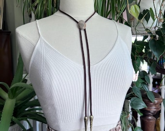 May include: A bolo tie with a dark brown cord and a circular, patterned stone centerpiece. The tie features decorative metal tips. The bolo tie is displayed on a white mannequin, with a white ribbed tank top and a plaid skirt.