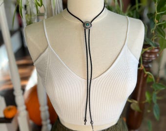 Puede incluir: Un corbatín bolo negro con una concha de plata y turquesa. El corbatín bolo se lleva sobre un top blanco sin mangas y pantalones verdes.