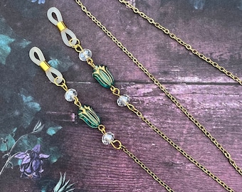 May include: A gold chain with clear crystal beads and green glass beads in the shape of a flower. The chain has two silicone loops at each end for attaching to glasses.