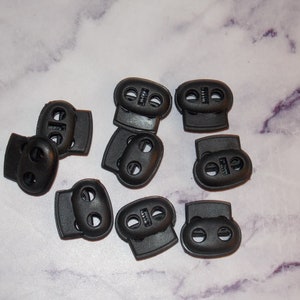 May include: Ten black plastic toggle clasps with two holes each. The clasps are oval shaped and have a smooth surface.
