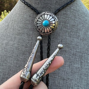 Pode incluir: Um la&ccedil;o bolo preto com um pingente de flor de prata e turquesa. O pingente tem um design detalhado e est&aacute; preso a um cord&atilde;o de couro tran&ccedil;ado.