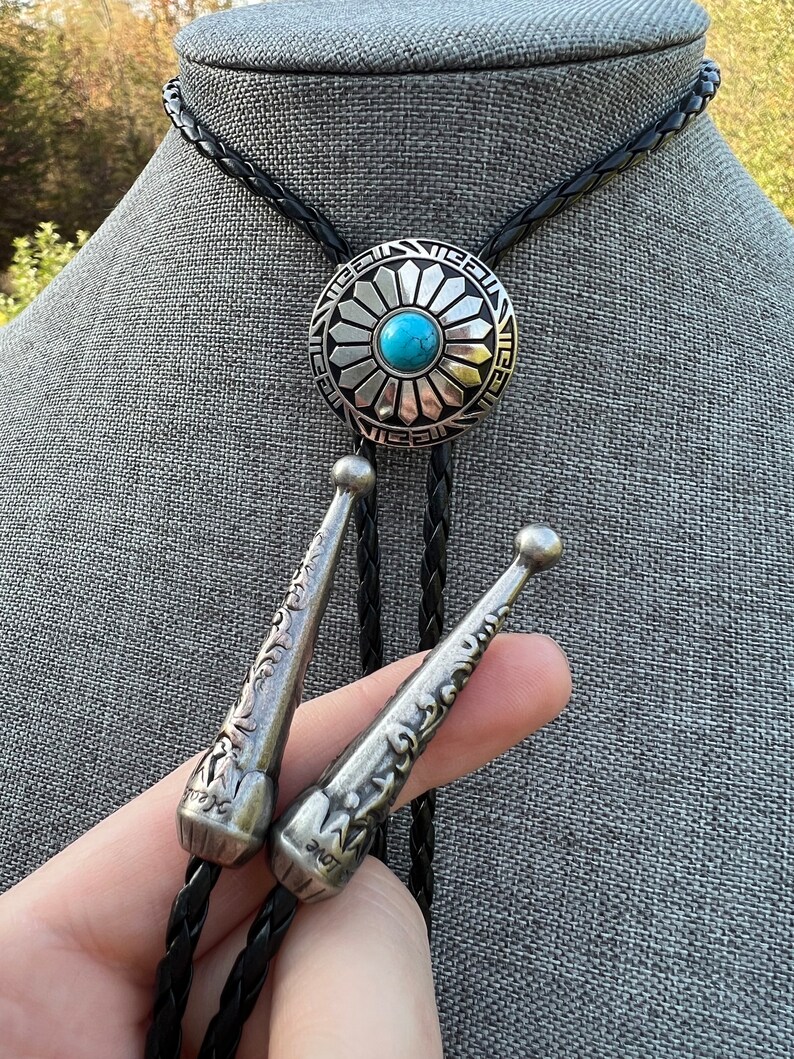 Pode incluir: Um la&ccedil;o bolo preto com um pingente de flor de prata e turquesa. O pingente tem um design detalhado e est&aacute; preso a um cord&atilde;o de couro tran&ccedil;ado.