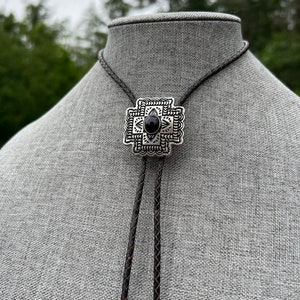 Op de afbeelding: Een zwarte bolo-das met een geometrische hanger van zilver en zwart. De hanger heeft een zwarte steen in het midden.