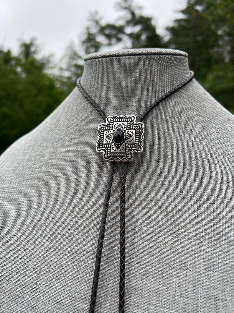 Op de afbeelding: Een zwarte bolo-das met een geometrische hanger van zilver en zwart. De hanger heeft een zwarte steen in het midden.