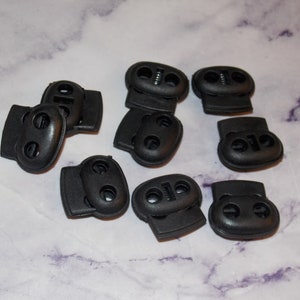 May include: A close-up of ten black plastic toggle clasps. The clasps have two holes and a spring mechanism.