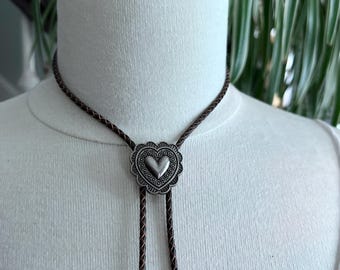 以下が含まれることがあります： ハート型のペンダントが付いたシルバーのボロタイ。ペンダントは装飾的な縁で囲まれ、茶色の編み込みコードから吊り下げられています。ボロタイは白いマネキンに飾られています。