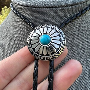 Pode incluir: Um la&ccedil;o bolo tran&ccedil;ado preto com um pingente de prata e turquesa com um design de sol estilizado.