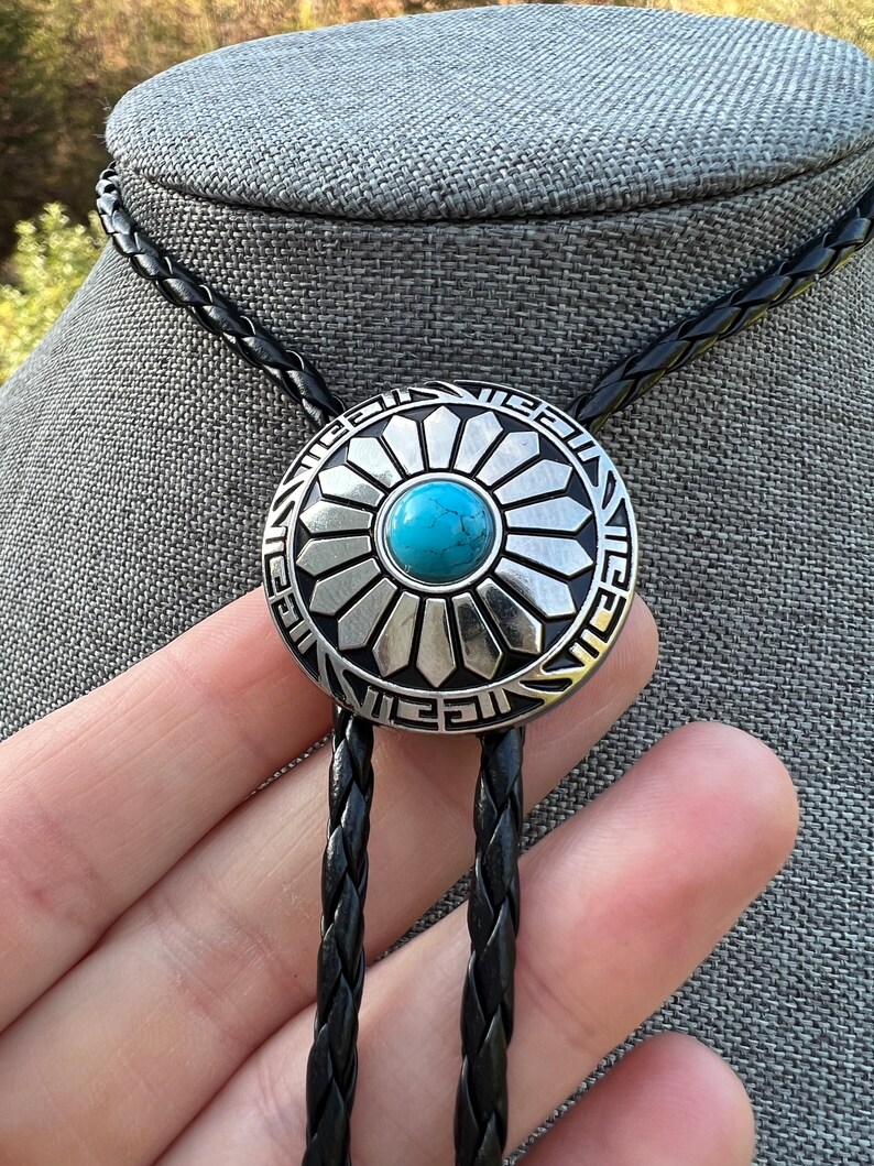 Pode incluir: Um la&ccedil;o bolo tran&ccedil;ado preto com um pingente de prata e turquesa com um design de sol estilizado.