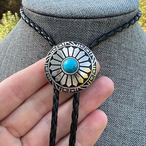 Pode incluir: Um bolo tie prateado com uma pedra turquesa no centro de um design circular. O bolo tie tem um cord&atilde;o tran&ccedil;ado preto.
