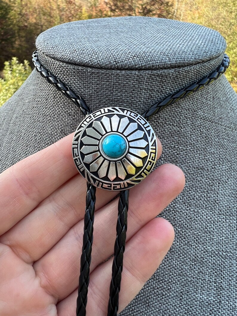 Pode incluir: Um bolo tie prateado com uma pedra turquesa no centro de um design circular. O bolo tie tem um cord&atilde;o tran&ccedil;ado preto.