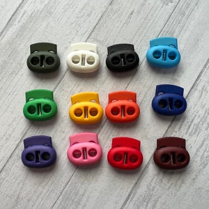 May include: Assortment of 12 plastic cord lock toggles in various colors, including green, white, black, blue, red, orange, yellow, purple, pink, brown, and dark blue.