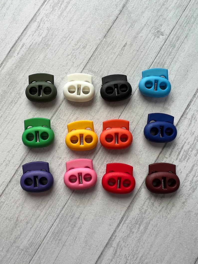 May include: Assortment of 12 plastic cord lock toggles in various colors, including green, white, black, blue, red, orange, yellow, purple, pink, brown, and dark blue.