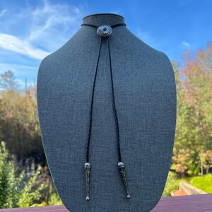 Pode incluir: Um la&ccedil;o bolo preto com uma concha de prata com uma pedra turquesa. A concha tem um design decorativo. O la&ccedil;o bolo &eacute; exibido em um manequim cinza.