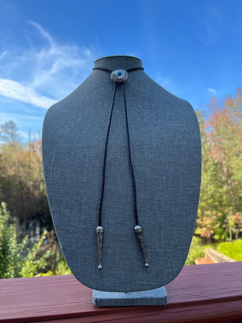 Pode incluir: Um la&ccedil;o bolo preto com uma concha de prata com uma pedra turquesa. A concha tem um design decorativo. O la&ccedil;o bolo &eacute; exibido em um manequim cinza.