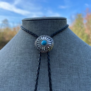 Pode incluir: Um cord&atilde;o bolo de couro preto com um pingente de prata e turquesa com um design circular com padr&otilde;es geom&eacute;tricos.