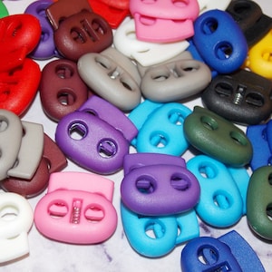 May include: Assortment of colorful plastic toggle clasps, also known as cord locks, in various colors including purple, blue, green, yellow, red, brown, white, and black.