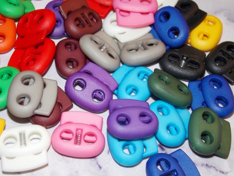 May include: Assortment of colorful plastic toggle clasps, also known as cord locks, in various colors including purple, blue, green, yellow, red, brown, white, and black.
