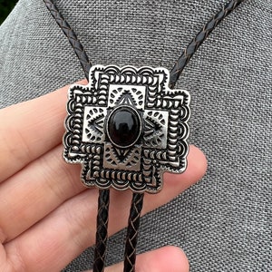 Op de afbeelding: Een zilveren bolo tie met een zwarte steen in het midden en een geometrisch ontwerp. De bolo tie is aan een zwart lederen koord.