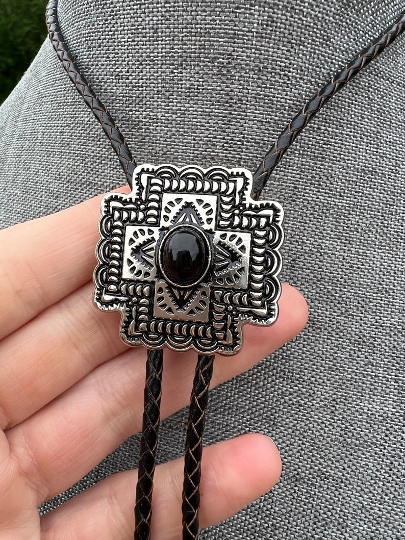 Op de afbeelding: Een zilveren bolo tie met een zwarte steen in het midden en een geometrisch ontwerp. De bolo tie is aan een zwart lederen koord.