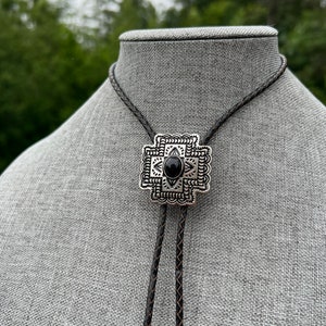 Op de afbeelding: Een zwarte bolo-das met een geometrische hanger van zilver en zwart. De hanger heeft een zwarte steen in het midden.
