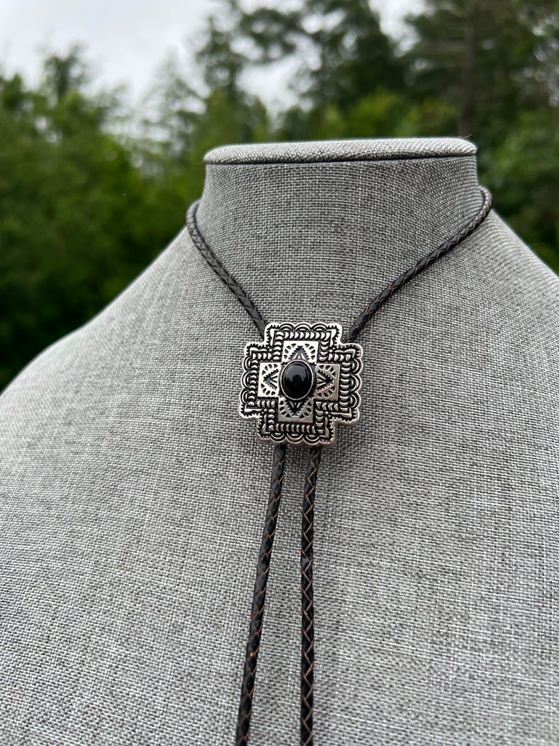 Op de afbeelding: Een zwarte bolo-das met een geometrische hanger van zilver en zwart. De hanger heeft een zwarte steen in het midden.