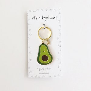 May include: A gold avocado keychain with a smiling face. The keychain is attached to a gold key ring. The card reads "it's a keychain!" and "a jar of pickles".