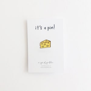 May include: A yellow enamel pin shaped like a smiling block of Swiss cheese with the text "it's a pin!" and "a jar of pickles. from the bay area, ca www.ajarofpickles.com"
