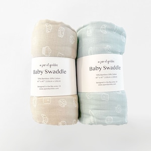 Puede incluir: Dos arrullos para bebé, uno en beige y otro en azul claro, con un estampado blanco de varios alimentos. Ambos arrullos están hechos de 70% bambú y 30% algodón y miden 120 cm por 120 cm. El texto "a jar of pickles Baby Swaddle" está impreso en la etiqueta de cada arrullo.
